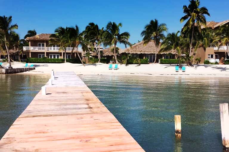 Wooden dock extending into calm turquoise water toward beachfront homes with palm trees