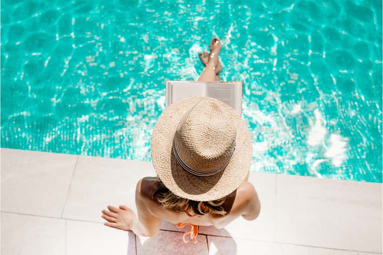 Woman in sunhat relaxing poolside, reading book with crystal-clear turquoise water behind her