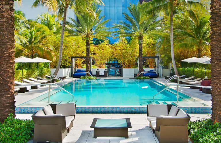 Luxury resort pool with turquoise water, palm trees, white umbrellas, and lounge chairs surrounded by modern architecture