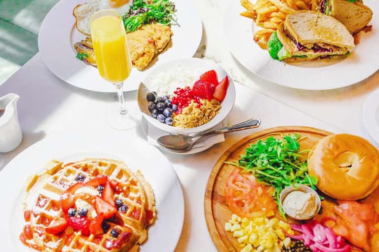 Brunch spread with waffles, pastries, fresh fruit bowl, juice, and sandwiches on white plates