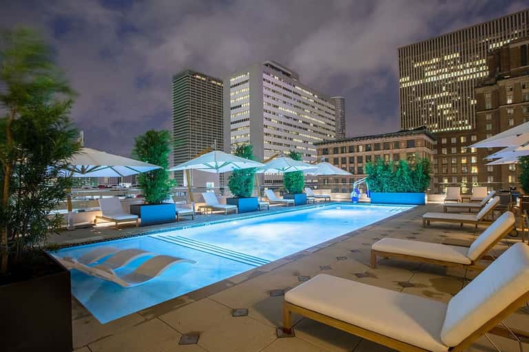 Rooftop pool illuminated at night with loungers, umbrellas, and downtown cityscape