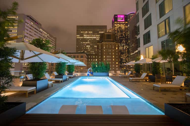 Illuminated rooftop pool at night with city skyline, lounge chairs, and umbrellas