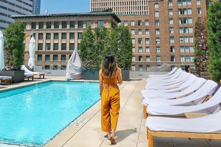 Woman in mustard dress standing at rooftop pool overlooking urban skyline with ivy-covered buildings