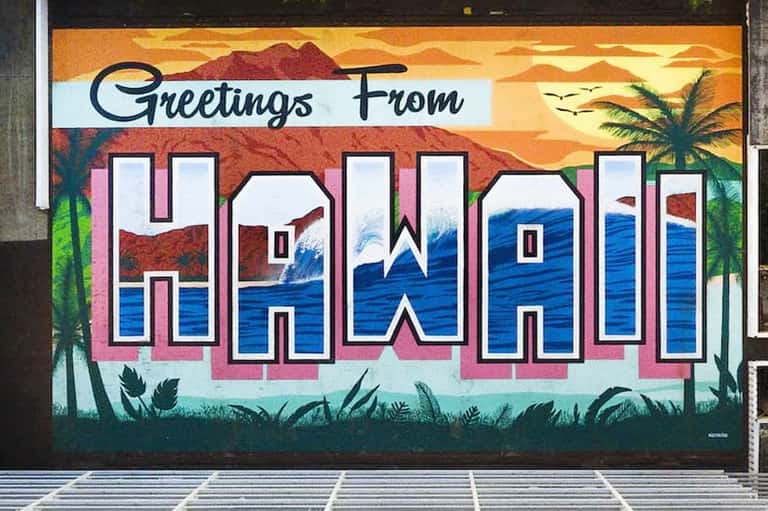 Vintage 'Greetings From Hawaii' mural with pink letters, ocean, mountains, and palm trees on building wall