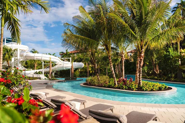 Tropical resort pool with waterslide, palm trees, lounge chairs, and vibrant red flowering plants