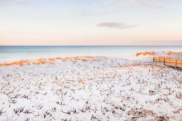 Serene beach at sunset with white sand, sea grass, wooden fence, and calm ocean waters