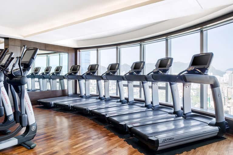 Modern gym with treadmills and exercise bikes lined along floor-to-ceiling windows overlooking a cityscape