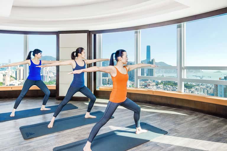 Three women in yoga class performing warrior pose with cityscape view through floor-to-ceiling windows
