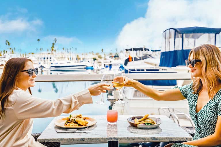Two women toasting wine glasses at waterfront restaurant overlooking marina