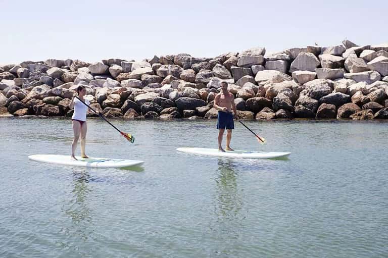 Two people paddleboarding on calm water near rocky breakwater