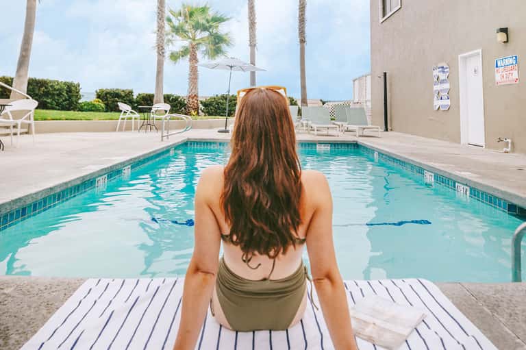 Woman in swimsuit sitting poolside at resort with palm trees and blue water