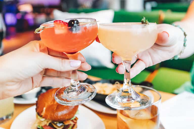 Two hands toasting with colorful cocktails at an outdoor event with food and decorations