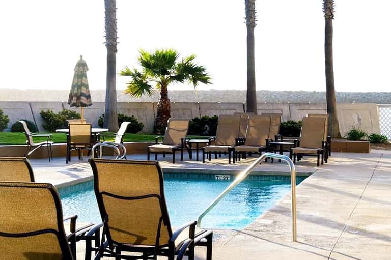 Poolside lounge area with tan chairs, umbrellas, palm trees, and mountain views in background