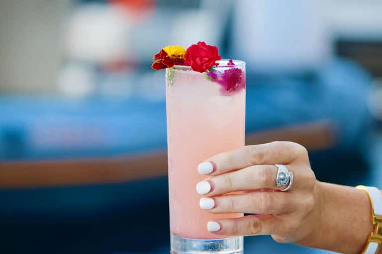 Pink cocktail with edible flowers garnish held by hand with ring, ocean backdrop