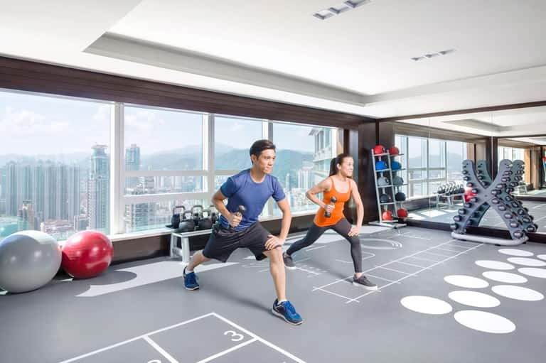 Two people exercising in a high-rise gym with floor-to-ceiling windows overlooking a cityscape and mountains