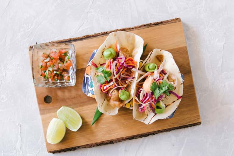 Gourmet tacos on wooden board with cilantro, pickled onions, lime wedges, and salsa