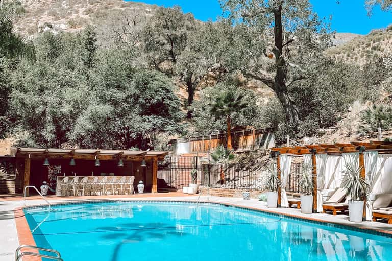 Resort pool surrounded by lounge chairs, wooden pergolas, and olive trees on hillside