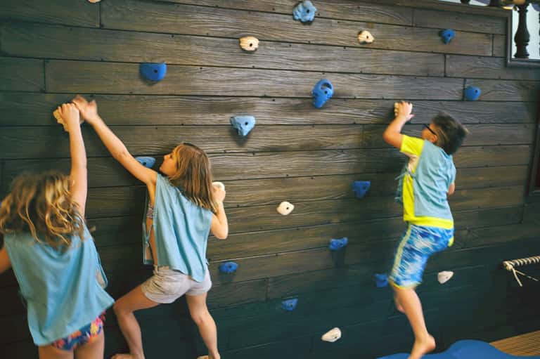 Children climbing a wooden wall with blue and white hand holds at an indoor activity center