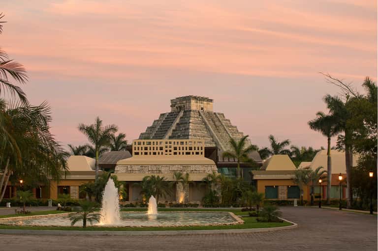 Mayan pyramid structure at dusk with fountains, palm trees, and warm architectural lighting