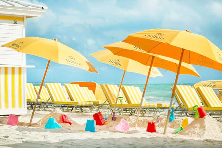 Beach scene with yellow and orange striped lounge chairs, Hilton branded umbrellas, colorful sand buckets, and turquoise ocean