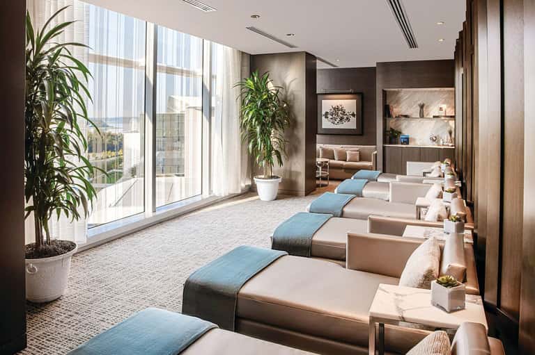 Modern spa relaxation room with beige lounge chairs, blue accents, plants, and floor-to-ceiling windows