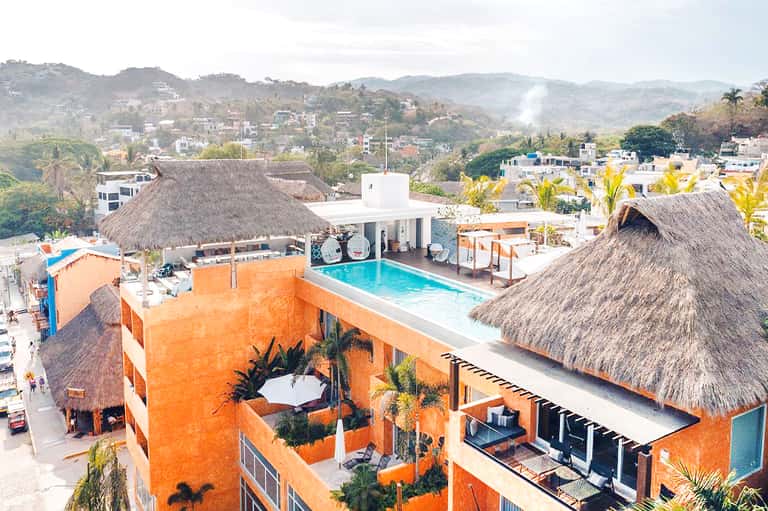 Aerial view of orange resort building with rooftop pool, thatched-roof structures, and hillside town landscape