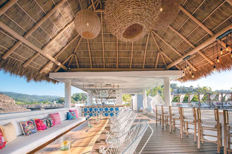 Beachfront tiki bar lounge with thatch roof, white woven chairs, and ocean views