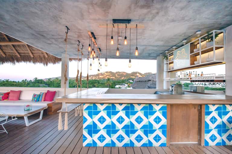 Modern beach bar with blue tile design, wooden counter, pendant lights, and ocean mountain views