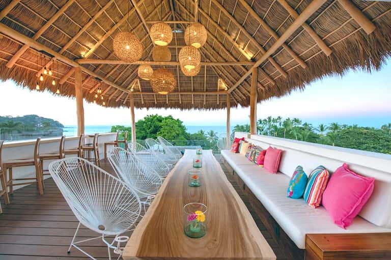 Covered beach pavilion with woven ceiling, wooden dining table, white chairs, and ocean view with palm trees