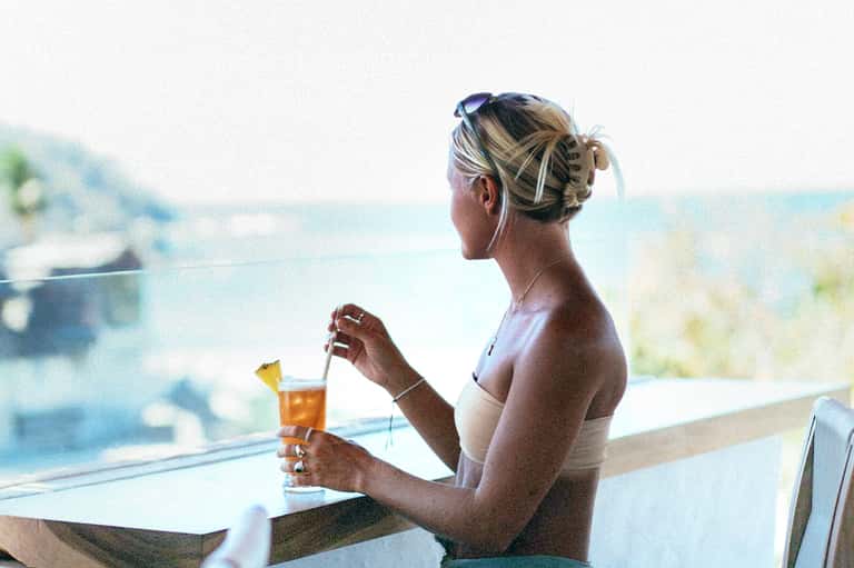 Woman in bikini enjoying a tropical drink while overlooking a scenic ocean view from balcony