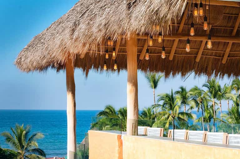Thatched-roof beach cabana with hanging lanterns overlooking ocean and palm trees