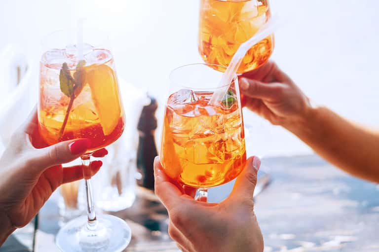 Hands holding tropical cocktails with ice and garnish against ocean and sky background