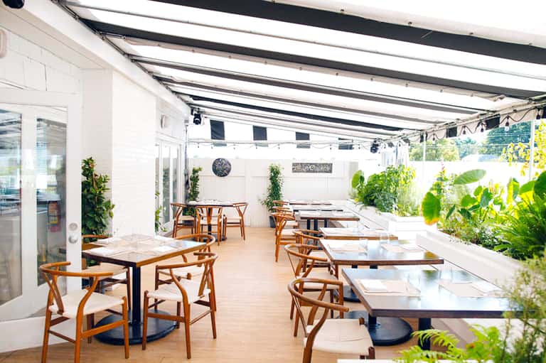 Modern restaurant patio with retractable awning, wooden chairs, and potted greenery