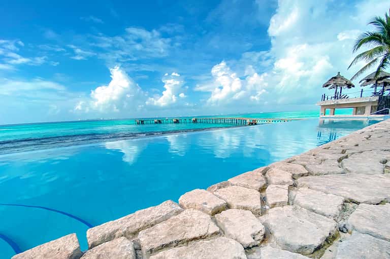 Turquoise infinity pool overlooking tropical ocean with pier, palm trees, and thatched umbrellas