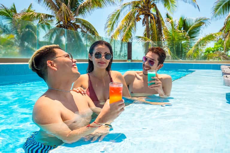 Three guests enjoying tropical drinks in a resort pool with palm trees