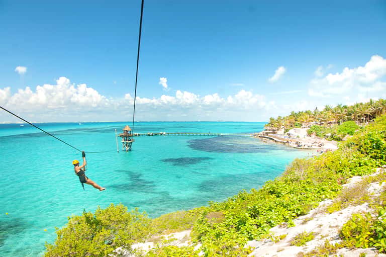Person zip-lining over turquoise ocean water with palm trees and beach resort visible below