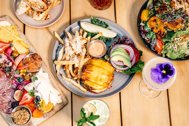Overhead view of gourmet plates with burgers, salads, charcuterie, and colorful cocktails on wood table