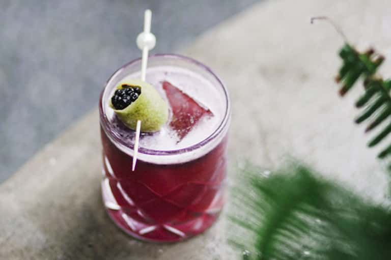 Colorful berry-garnished cocktail with purple liquid on concrete surface with blurred greenery background