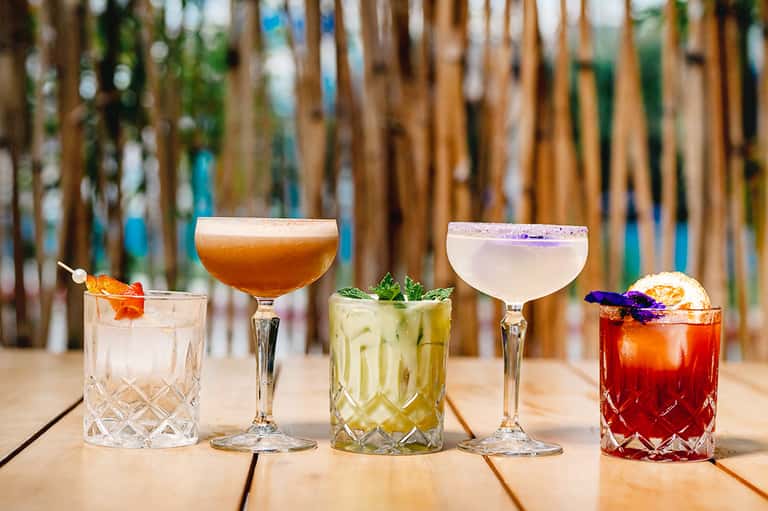 Five colorful cocktails on wooden deck with beach fence background and tropical garnishes