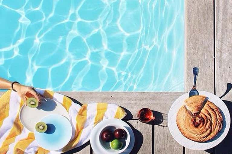 Poolside breakfast spread with pastry, fruit, and beverages on wooden deck next to clear blue water