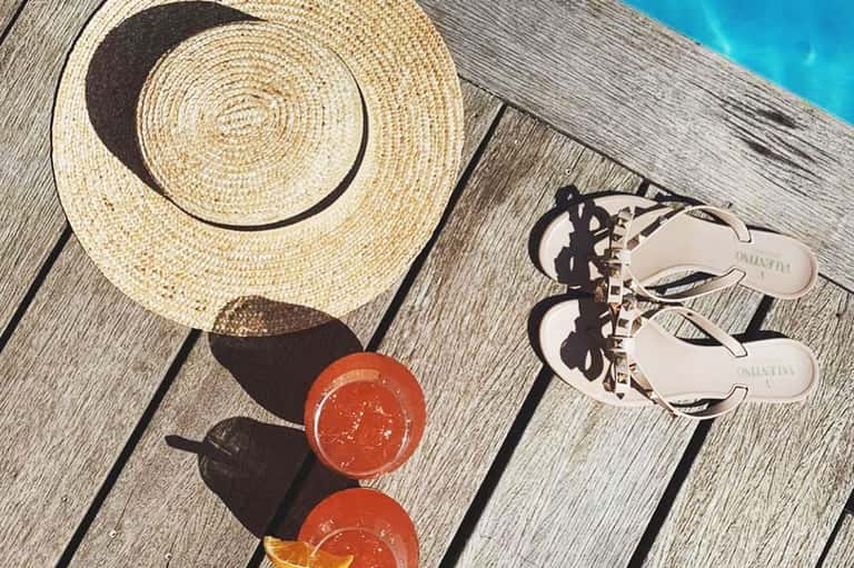 Beach accessories on wooden dock: straw hat, white sandals, and coral-colored cocktails with ocean and sky backdrop