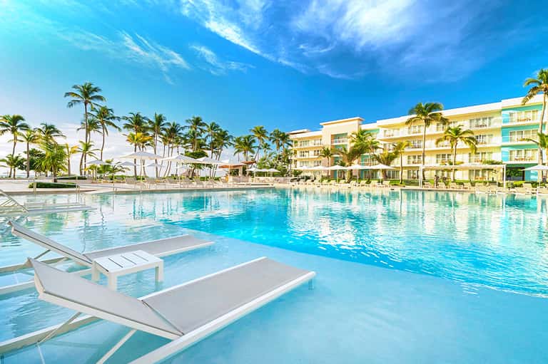 Resort pool with lounge chairs, palm trees, and pastel-colored hotel buildings under blue sky