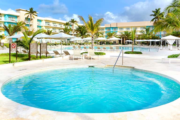 Beachfront resort pool area with palm trees, umbrellas, and modern hotel buildings