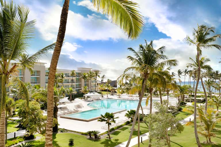 Resort pool surrounded by palm trees with modern multi-story hotel building and ocean view