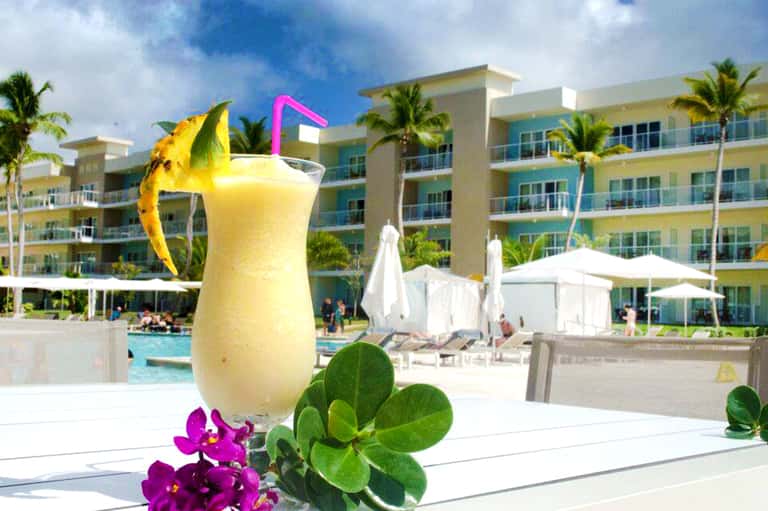 Tropical resort poolside with yellow cocktail, colorful buildings, palm trees, and white umbrellas