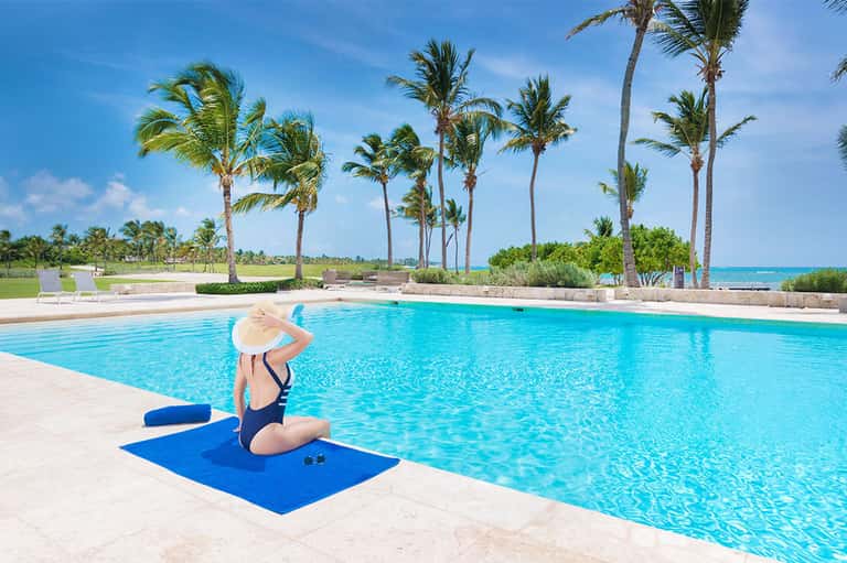Woman in hat sitting poolside with palm trees and ocean beyond