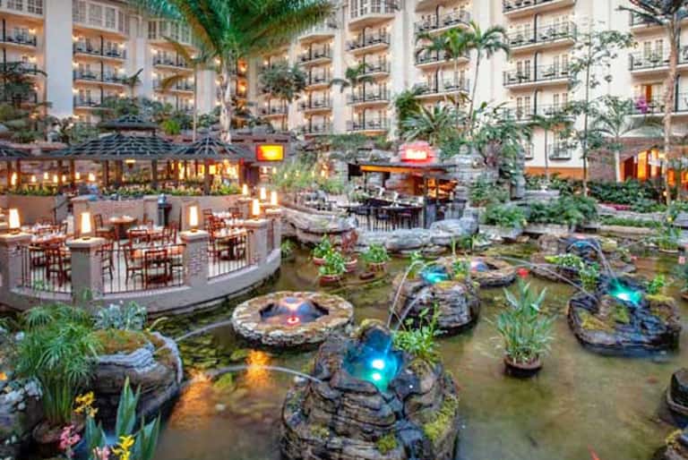 Indoor atrium with dining areas, koi pond with lit rocks, tropical plants, and multi-story hotel buildings surrounding the space