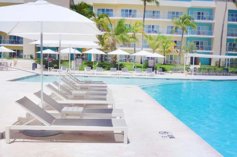 Resort pool with white lounge chairs and umbrellas, flanked by palm trees and colorful building