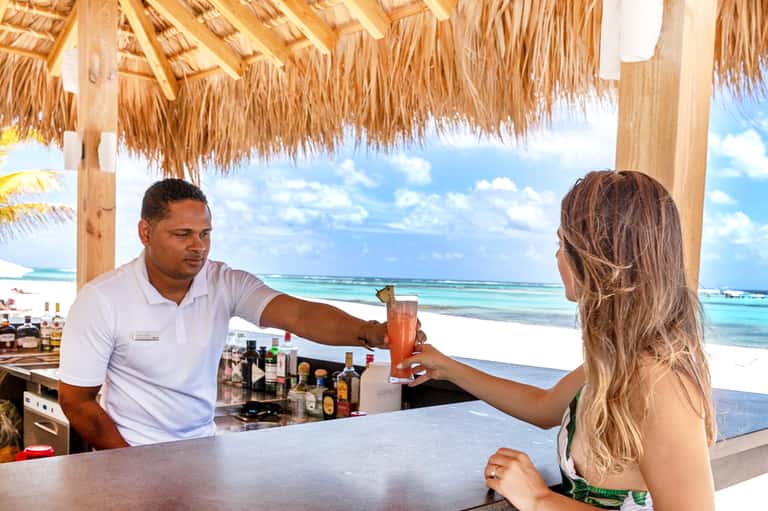 Beach bartender serving a coral-colored cocktail to a guest under a thatched-roof beach bar