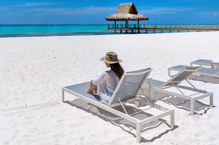 Woman relaxing on beach lounge chair facing turquoise ocean with thatched pier structure